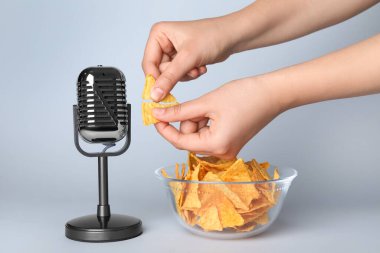 Gri arka planda mikrofon ve nacho çipli ASMR sesleri çıkaran kadın, yakın plan.