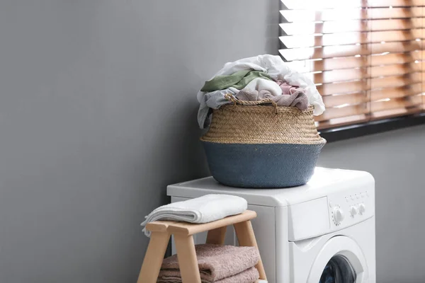 Wicker basket with dirty laundry on washing machine near grey wall indoors