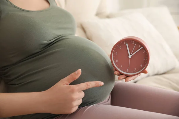 Young pregnant woman pointing at clock near her belly indoors, closeup. Time to give birth