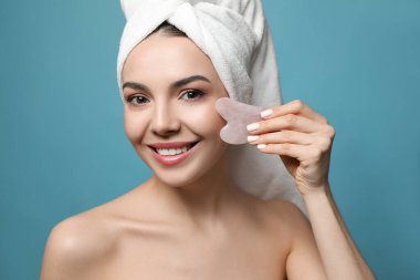 Güzel genç bir kadın mavi arka planda gua sha aracıyla yüz masajı yapıyor.