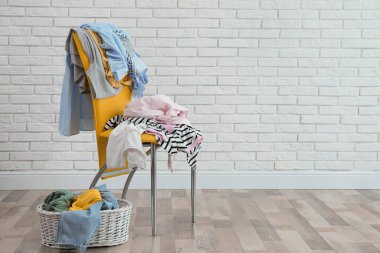 Beyaz tuğla duvarın yanında farklı kıyafetlerle sarı sandalye ve hasır sepet.