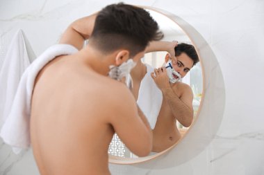 Yakışıklı genç adam banyoda aynanın yanında tıraş oluyor.