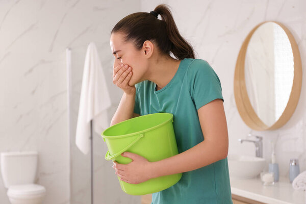 Молодая женщина с ведром страдает от тошноты в ванной. Продовольственное отравление