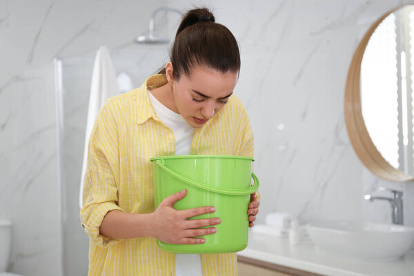 Молодая женщина с ведром страдает от тошноты в ванной. Продовольственное отравление