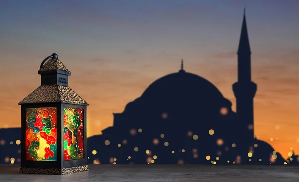 Decorative Arabic lantern on stone surface and silhouette of mosque at sunset on background, space for text