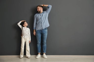 Father and daughter comparing their heights near black wall indoors, space for text clipart