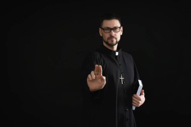 Priest with Bible making blessing gesture on dark background, space for text clipart