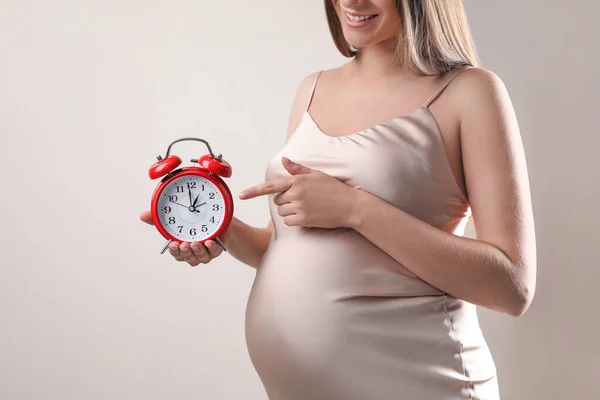 ベージュを背景に腹近くの目覚まし時計を指差す若い妊婦さんが近づいています 出産までの時間 — ストック写真