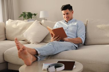 Oturma odasındaki kanepede kitap okuyan adam.