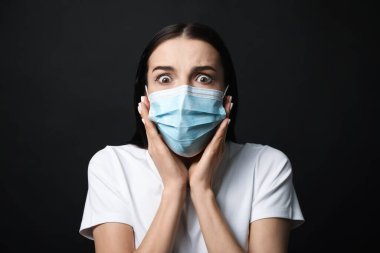 Koruyucu maskeli genç bir kadın siyah arka planda korku hissediyor.