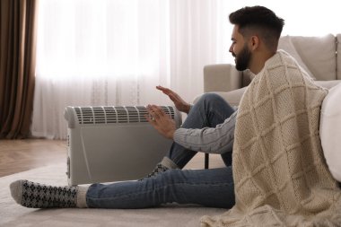 Genç adam elektrikli ısıtıcının yanında ellerini ısıtıyor.
