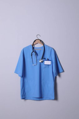 Açık gri arka planda asılı steteskop, rozet ve kalemli doktor gömleği.