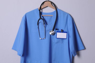 Açık gri arka planda asılı steteskop, rozet ve kalemli doktor gömleği.