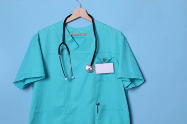 Açık mavi arkaplanda steteskop ve rozetli turkuaz doktor gömleği asılı.