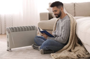 Genç adam elektrikli ısıtıcının yanında ısınıyor.