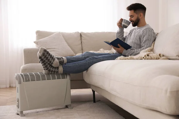 Junger Mann Mit Buch Und Heißgetränk Wärmt Füße Auf Elektroheizung — Stockfoto