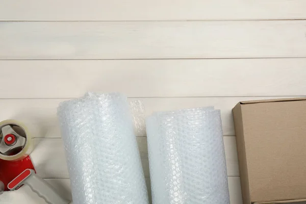 Bubble wrap rolls, cardboard box and tape dispenser on white wooden background, flat lay. Space for text