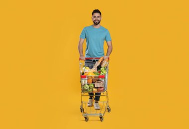 Sarı arka planda alışveriş arabası dolu mutlu bir adam.