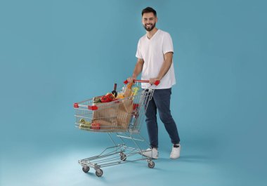 Açık mavi arka planda alışveriş arabalı mutlu bir adam.
