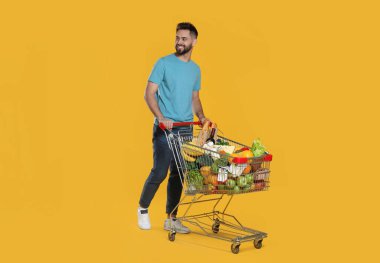 Sarı arka planda alışveriş arabası dolu mutlu bir adam.