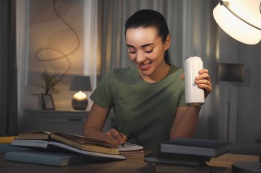Enerji içeceği içen genç bir kadın evde ders çalışıyor.
