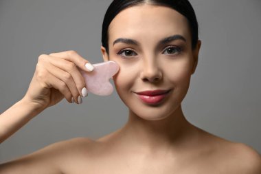 Beautiful young woman doing facial massage with gua sha tool on grey background, closeup clipart