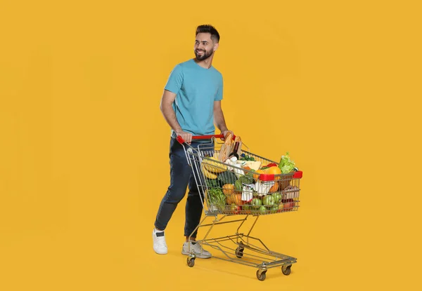 Glücklicher Mann Mit Einkaufswagen Voller Lebensmittel Auf Gelbem Hintergrund — Stockfoto
