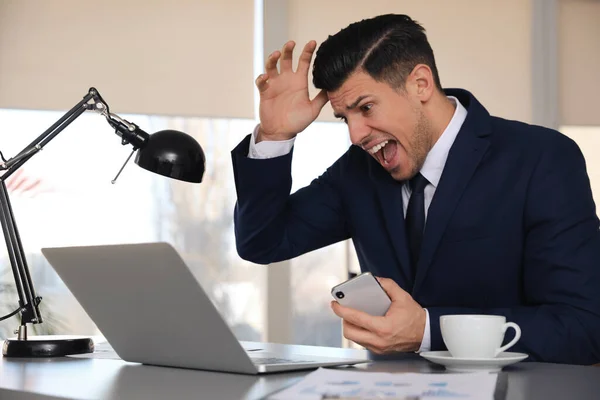 オフィスのテーブルでノートパソコンやスマートフォンを持つ感情的なビジネスマン — ストック写真