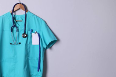 Turkuaz doktor gömleği, steteskop, rozet ve açık gri arka planda asılı kalem. Metin için boşluk