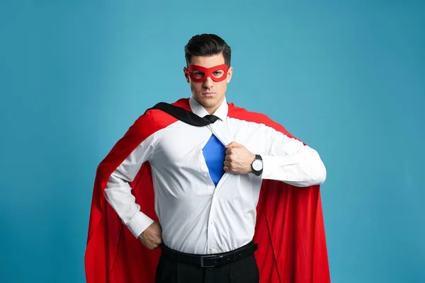 Businessman in superhero cape and mask taking shirt off on light blue background