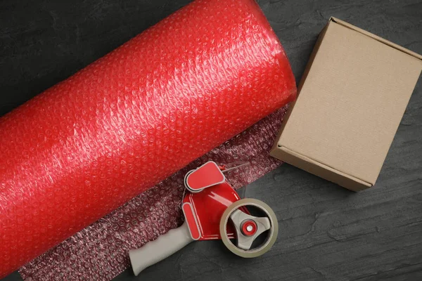 Bubble wrap roll, cardboard box and tape dispenser on black background, flat lay