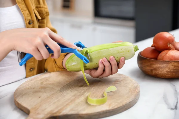 Woman peeling zucchini at table in kitchen, closeup. Preparing vegetable