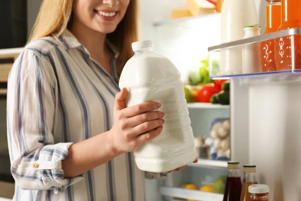 Young woman with gallon of milk near refrigerator in kitchen, closeup
