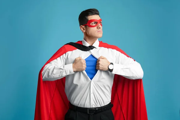 Businessman in superhero cape and mask taking shirt off on light blue background