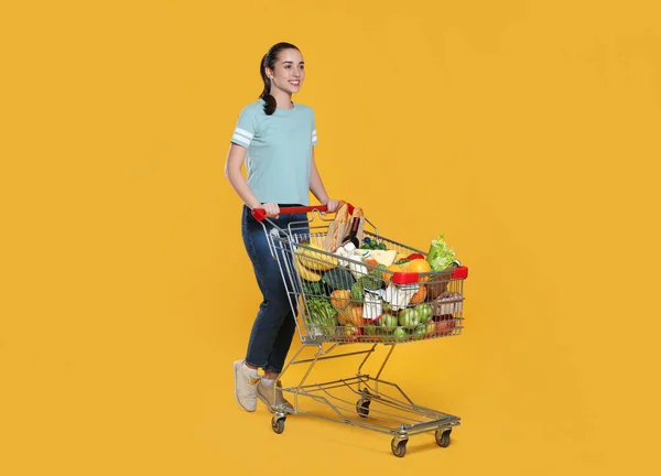 Glückliche Frau Mit Einkaufswagen Voller Lebensmittel Auf Gelbem Hintergrund — Stockfoto