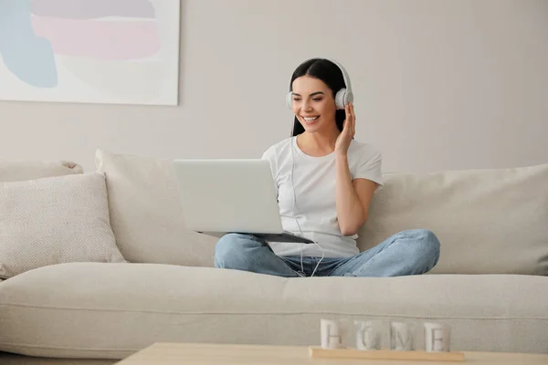 ノートパソコンとヘッドフォンを持つ女性は自宅でソファに座って — ストック写真