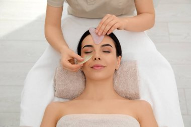 Genç bir kadın güzellik salonunda gua sha aracıyla yüz masajı yaptırıyor.