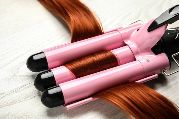 Curling iron with red hair lock on white wooden table, closeup