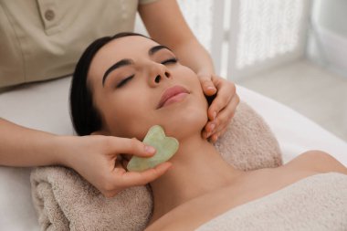 Genç bir kadın güzellik salonunda gua sha aracıyla yüz masajı yaptırıyor.