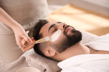 Kozmetik uzmanı spa salonunda adamın yüzüne maske takıyor.