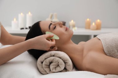 Young woman receiving facial massage with gua sha tool in beauty salon clipart