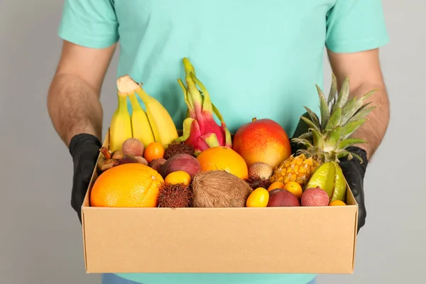 Courier holding box with assortment of exotic fruits on grey background, closeup