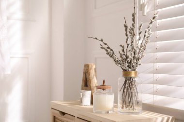 Glass vase with pussy willow tree branches and candle near window in room, space for text
