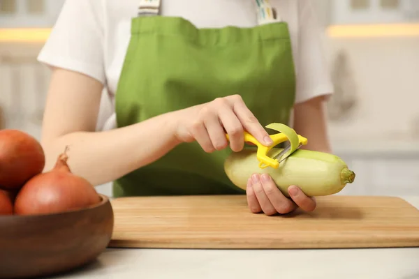 Woman peeling zucchini at table in kitchen, closeup. Preparing vegetable