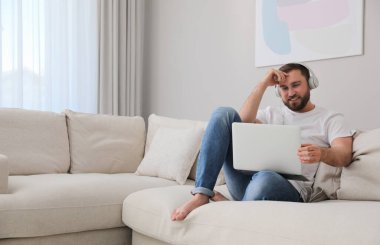 Dizüstü bilgisayarı ve kulaklığı olan bir adam evdeki koltukta oturuyor.