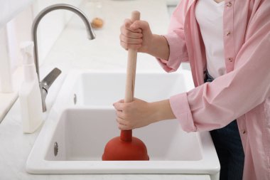 Woman using plunger to unclog sink drain in kitchen, closeup clipart