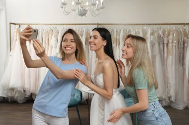 Gelinle fotoğraf çektiren arkadaşlar butikte gelinlik giyiyorlar.