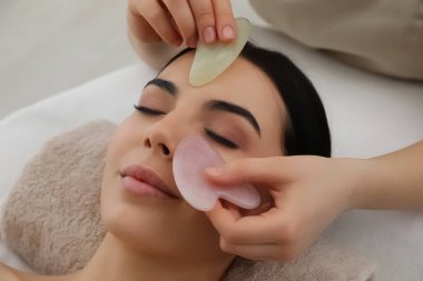 Genç bir kadın güzellik salonunda gua sha aletleriyle yüz masajı yaptırıyor.