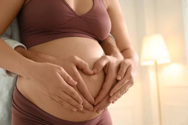 妊娠中の若い女性は彼女の近くの自宅で腹と夫に手で心を作って クローズアップ — ストック写真