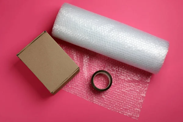 Bubble wrap roll, cardboard box and tape on pink background, flat lay